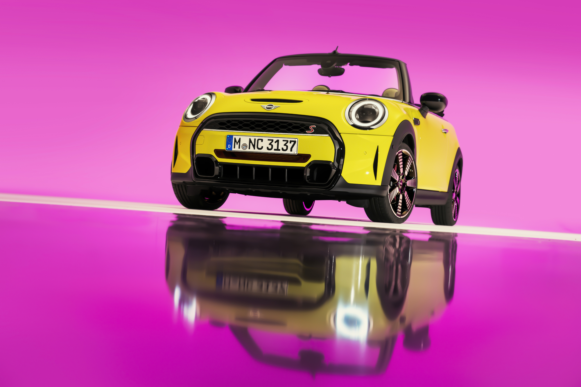 A yellow 2022 Mini Cooper S Convertible on display with a pink background