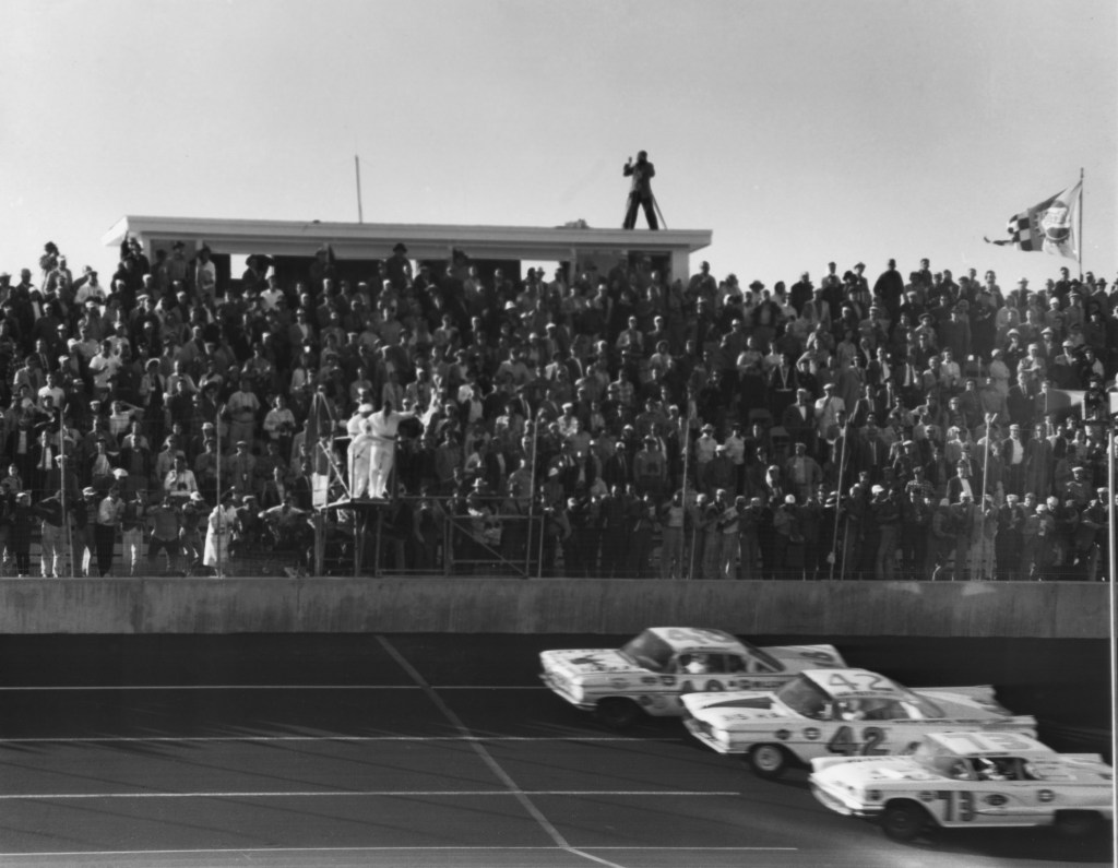 Three NASCAR cars, including Lee Petty's No. 42 Oldsmobile Super 88, head toward the finish line at the first Daytona 500 in 1959
