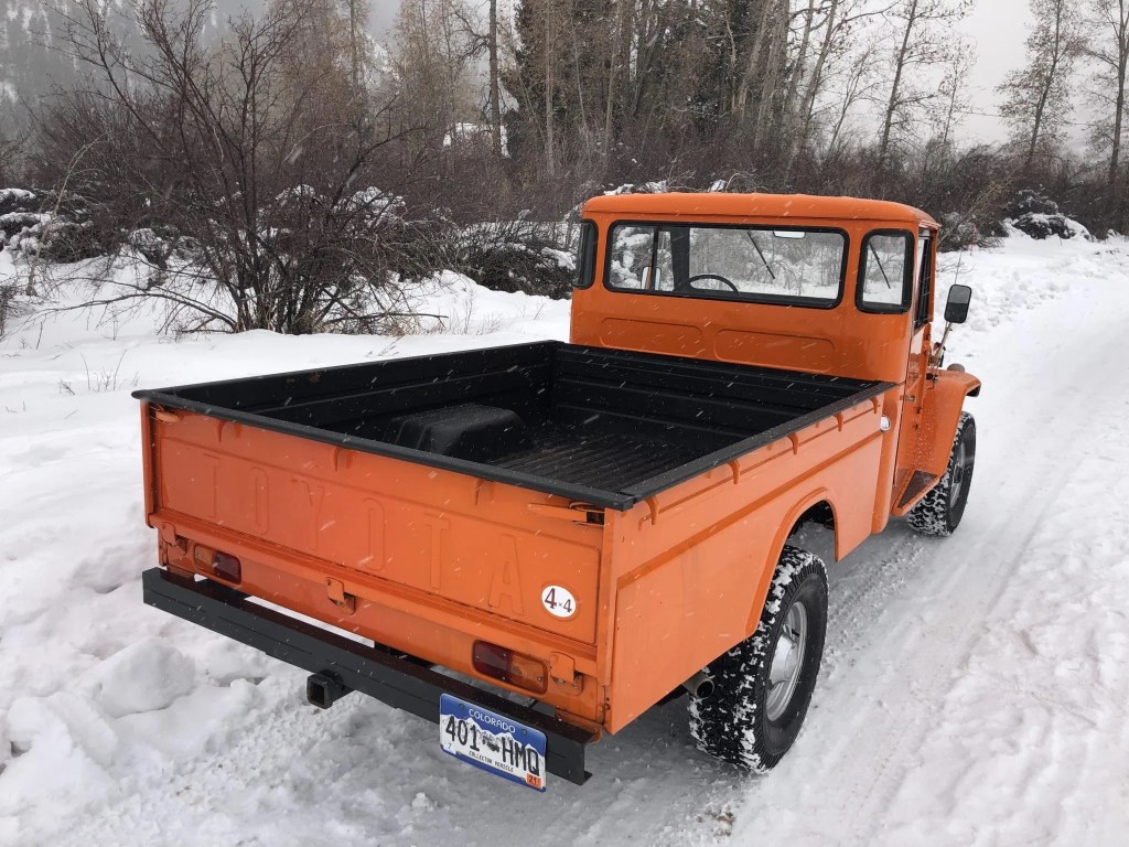 The rear 3/4 view of an orange 1964 FJ45 Toyota Land Cruiser pickup on a snowy road