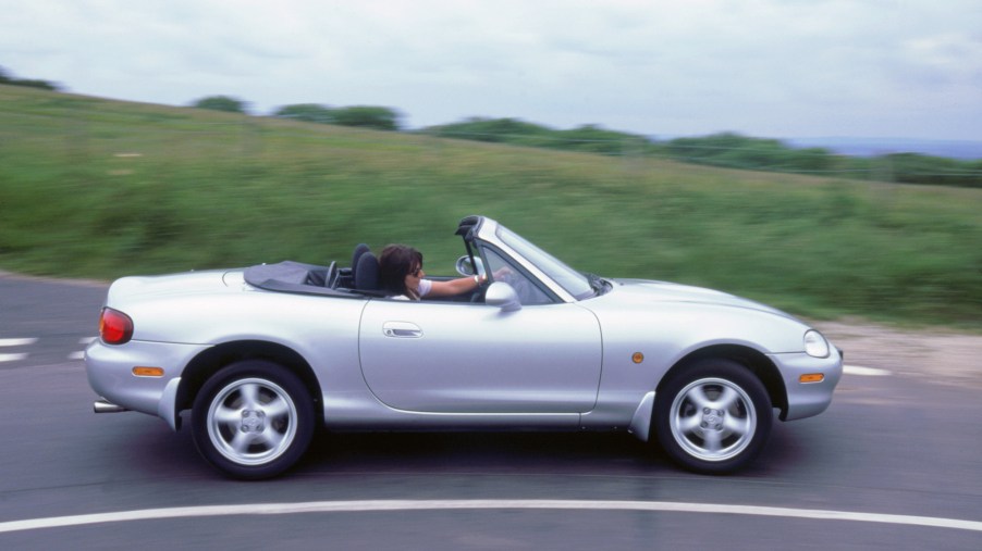 A person drives a silver 1999 Mazda MX5 on a curve in a road