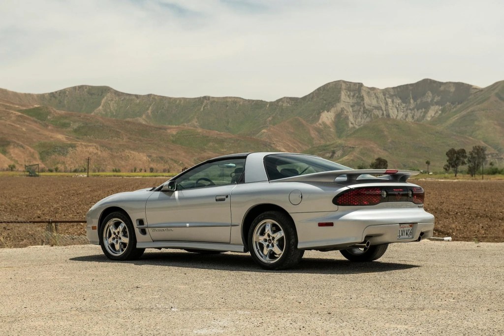 The rear 3/4 view of a silver 2002 Pontiac Firebird Trans Am WS6 on a dirt parking lot by a mountain range
