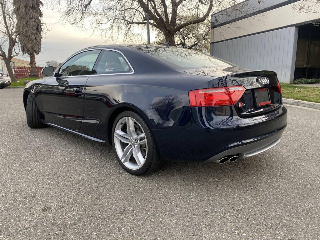 The rear 3/4 view of a dark-blue 2009 Audi S5 Coupe parked in a lot by a warehouse