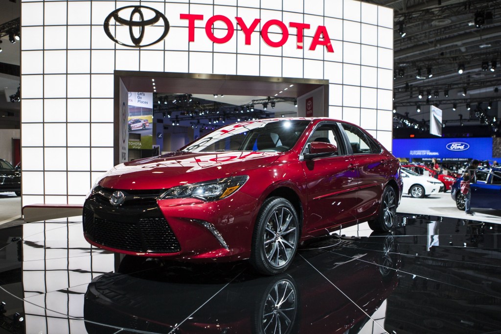 A red 2015 Toyota Camry on display at an auto show with Toyota in bold letters on the wall behind it.