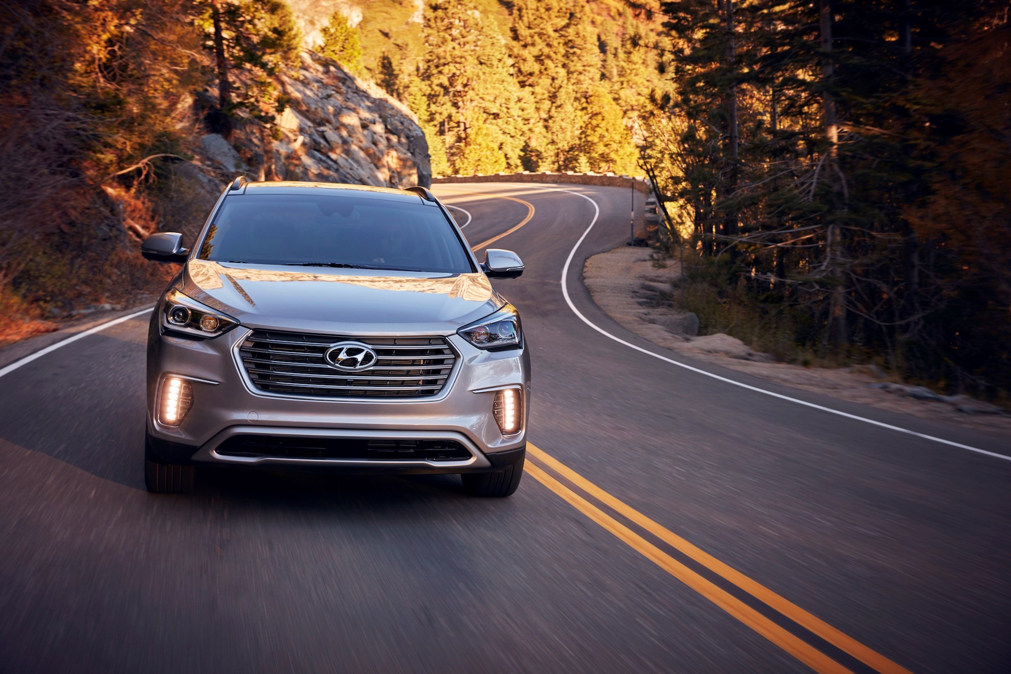 A silver 2018 Hyundai Santa Fe travels on a curvy two-lane mountain highway flanked by pine trees