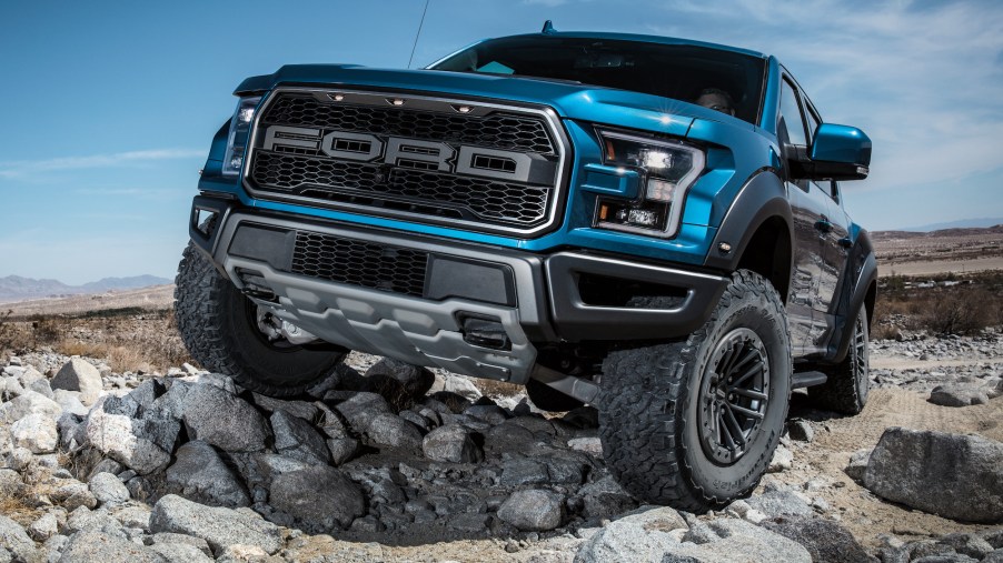 A bright-blue 2020 Ford F-150 Raptor parked on large rocks in a wide-open space with mountains and a blue sky in the background