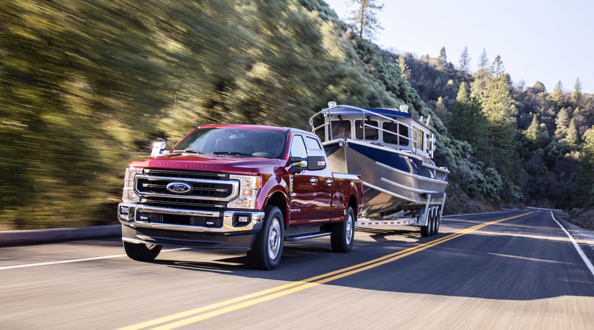 A red 2020 Ford F-250 King Ranch four-door diesel pickup truck towing a boat on a two-lane highway flanked by hills and pine trees