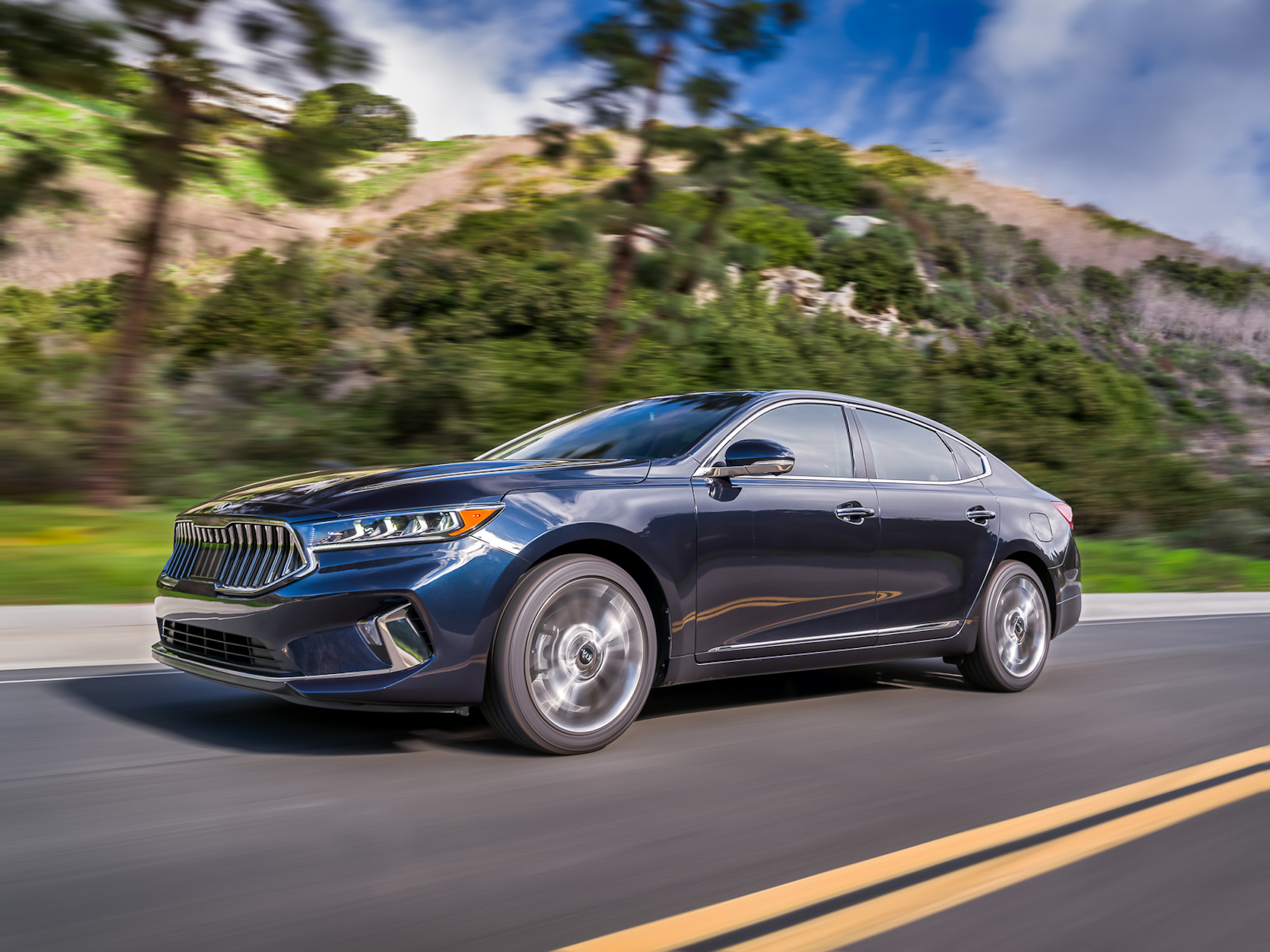 A dark-blue 2020 Kia Cadenza travels on a two-lane highway lined with pine trees and mountains