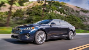 A dark-blue 2020 Kia Cadenza travels on a two-lane highway lined with pine trees and mountains