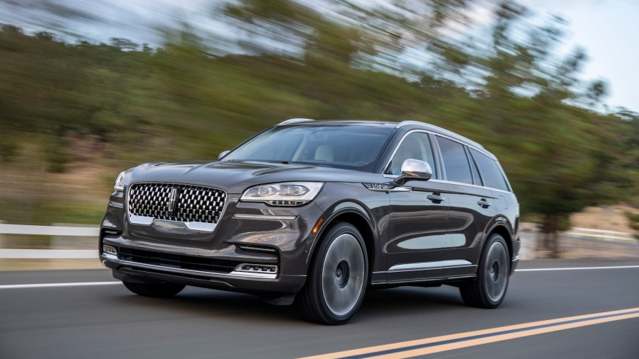 A dark-gray 2020 Lincoln Aviator travels on a two-lane highway along a white fence and trees