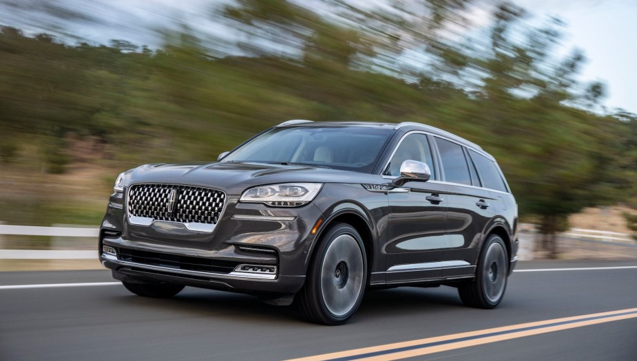 A dark-gray 2020 Lincoln Aviator travels on a two-lane highway along a white fence and trees