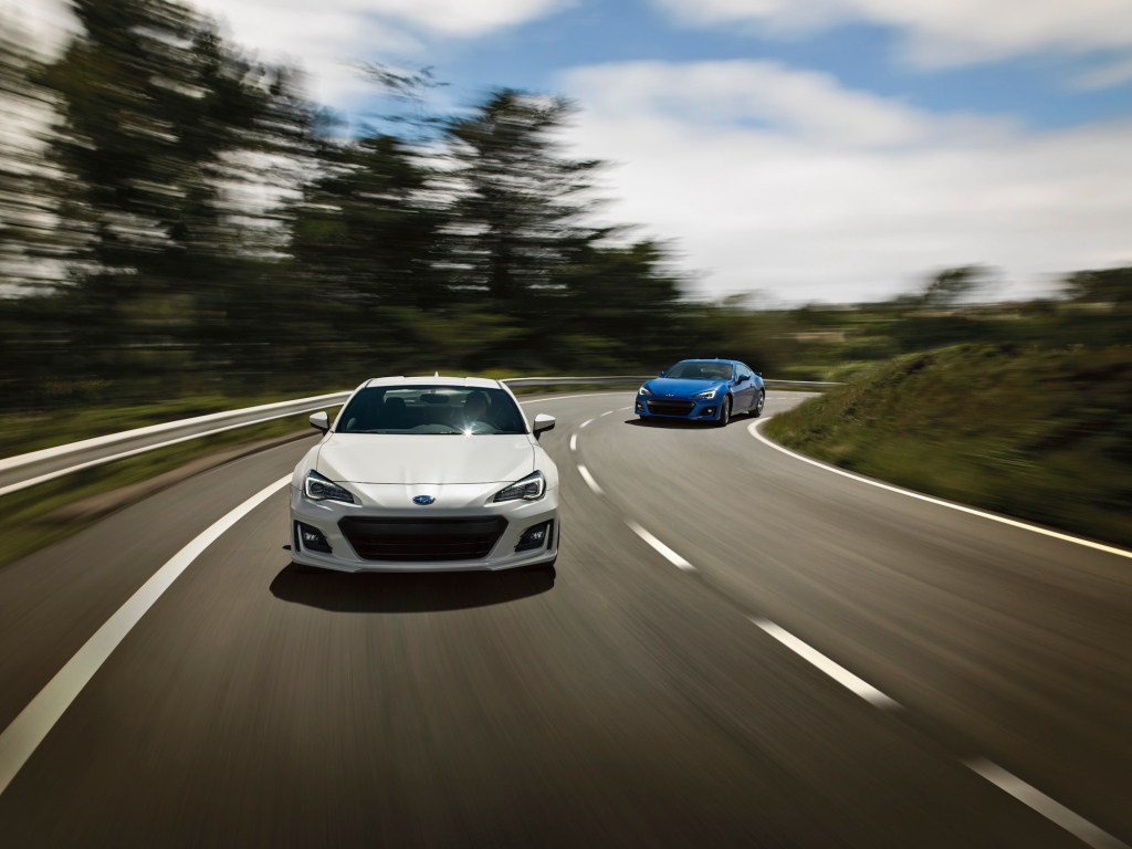 A white and a blue 2020 Subaru BRZ race on a winding two-lane highway flanked by hills and trees