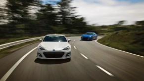 A white and a blue 2020 Subaru BRZ race on a winding two-lane highway flanked by hills and trees