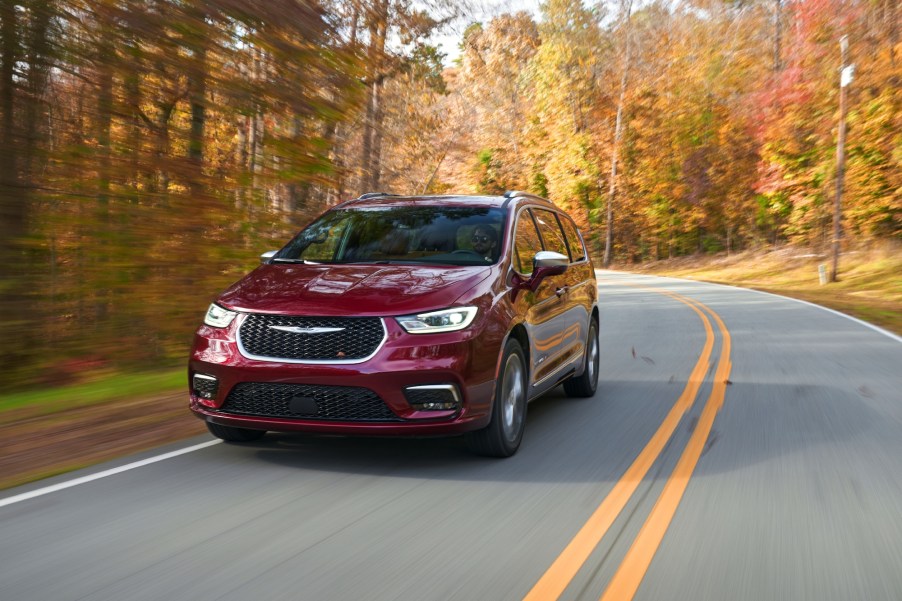 A red all-wheel-drive 2021 Chrysler Pacifica Pinnacle minivan traveling on a two-lane highway flanked by trees with autumn leaves