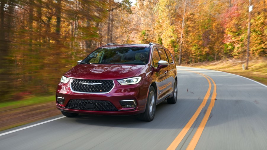 A red all-wheel-drive 2021 Chrysler Pacifica Pinnacle minivan traveling on a two-lane highway flanked by trees with autumn leaves