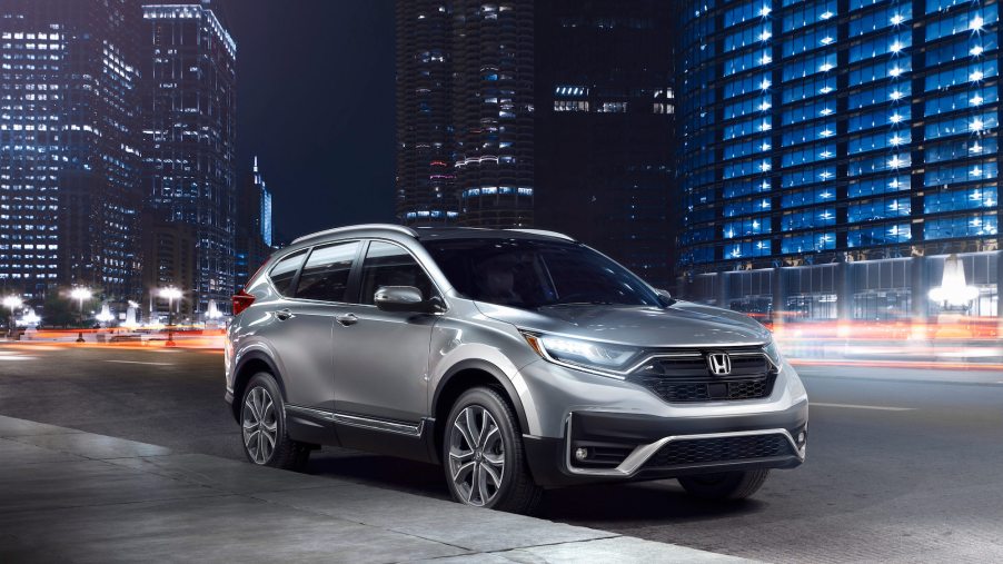 A silver 2021 Honda CR-V Touring model with its headlights on while parked on a city street at night