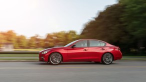 A red 2021 Infiniti Q50 sedan travels on a two-lane road alongside a park and trees