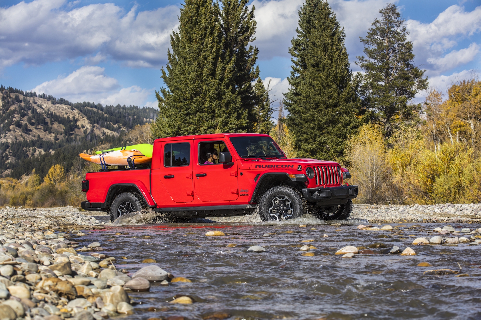 A red 2021 Jeep Gladiator Rubicon pickup truck carries kayaks in its bed while navigating a stream surrounded by pine trees and mountains