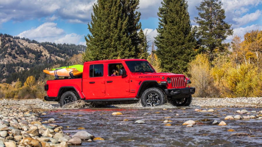 A red 2021 Jeep Gladiator Rubicon pickup truck carries kayaks in its bed while navigating a stream surrounded by pine trees and mountains