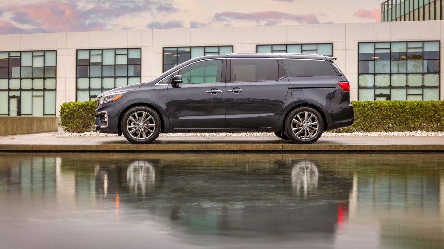 A dark-gray 2021 Kia Sedona parked between a body of water and a low-slung modern building