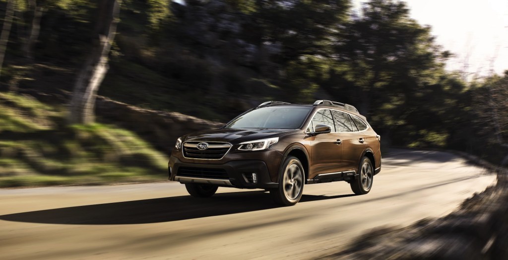 A brown 2021 Subaru Outback travels on a rural road flanked by trees and green hills