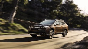 A brown 2021 Subaru Outback travels on a rural road flanked by trees and green hills