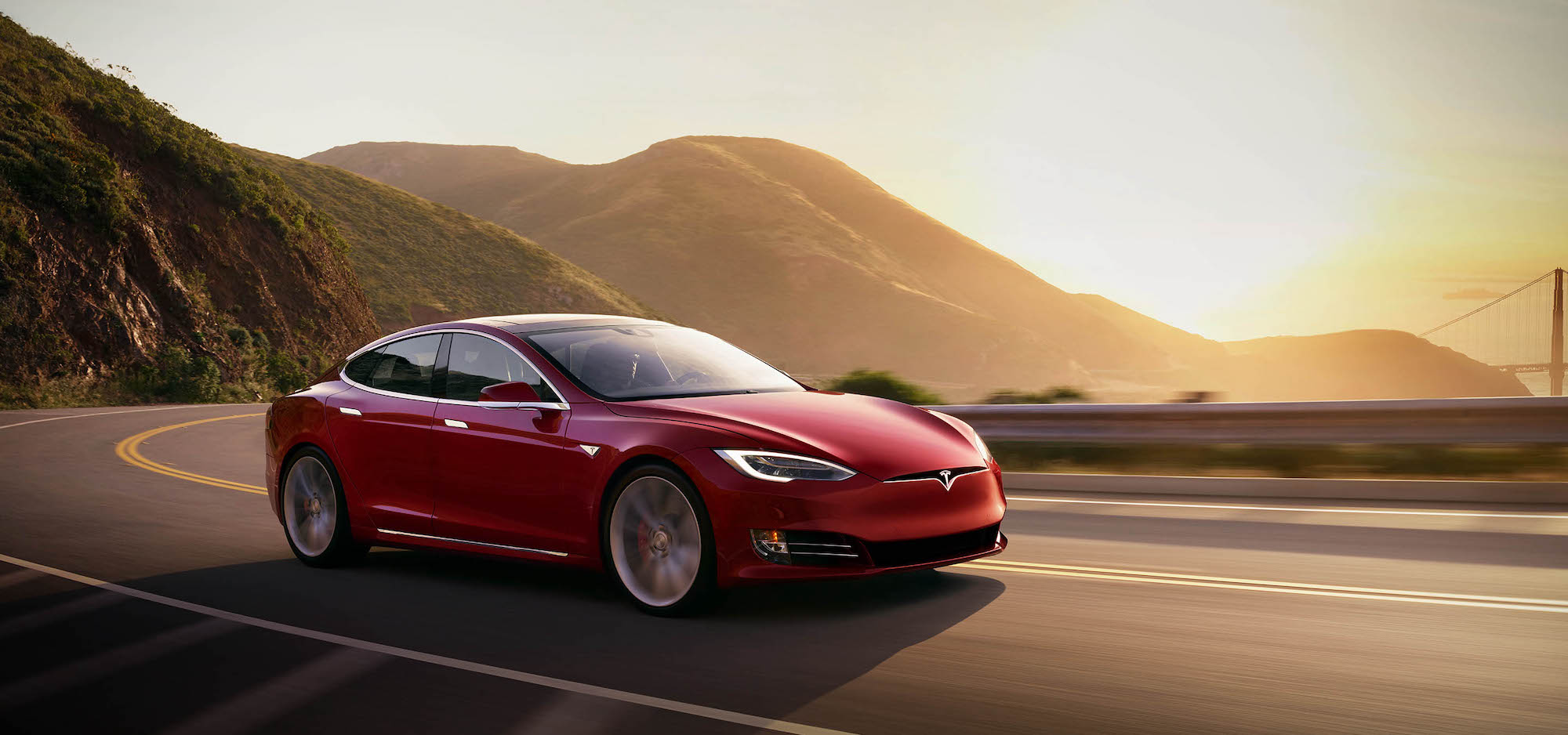 A red 2021 Tesla Model S travels on a two-lane highway in the mountains overlooking a body of water at sunset