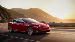 A red 2021 Tesla Model S travels on a two-lane highway in the mountains overlooking a body of water at sunset