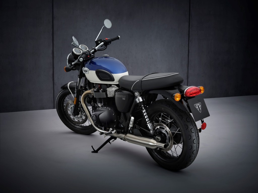 The rear 3/4 view of a blue-and-white 2022 Triumph Bonneville T100 in front of a gray background