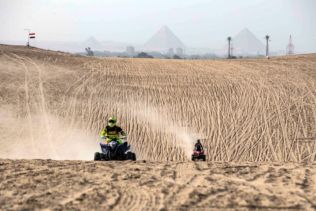 two riders going head to head in an ATV race in the sahara desert with egyptian pyramids in the background