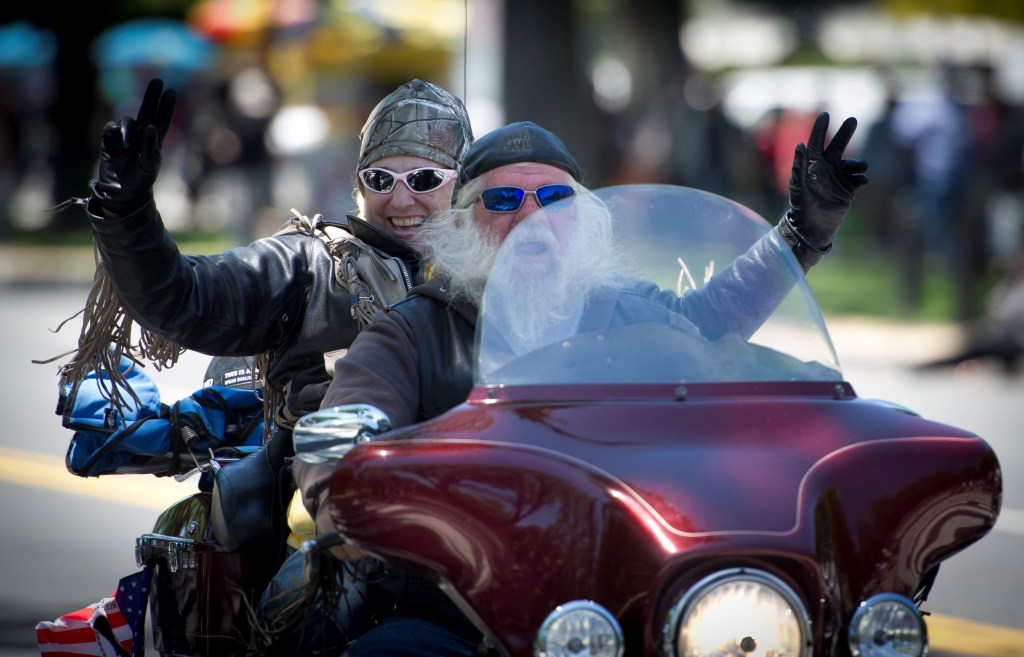 A motorcycle rider and passenger wave from a maroon Harley-Davidson