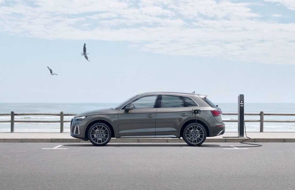 A side image of a 2021 Audi Q5 55 TFSI e quattro  in Daytona grey plugged into a hybrid charging station along the water