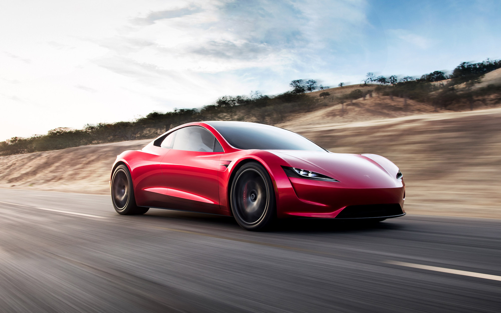 A red Tesla Roadster travels on a highway along brown hills dotted with trees