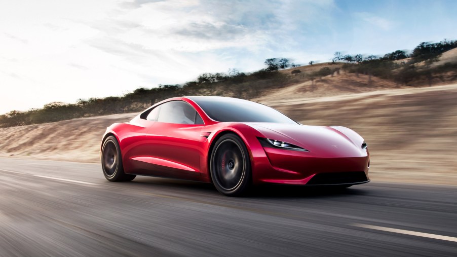 A red Tesla Roadster travels on a highway along brown hills dotted with trees