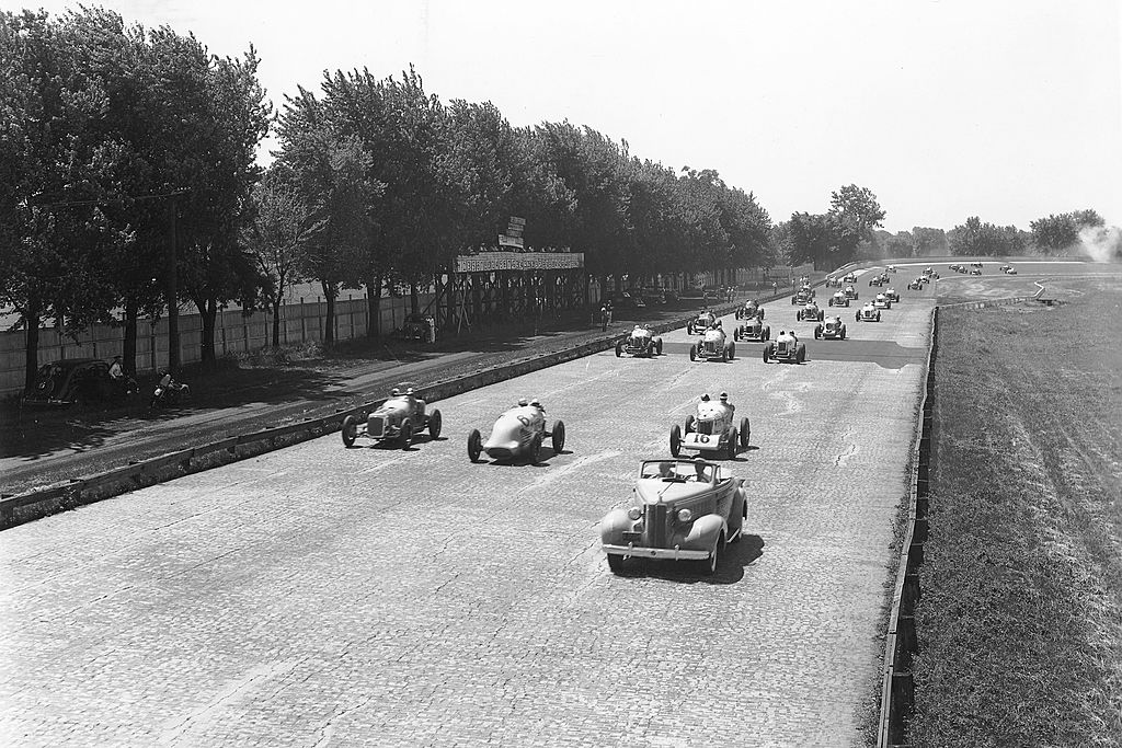 Indy cars are shown on the brick racing surface as drivers make their way around the pace lap of the Indianapolis 500 at the Indianapolis Motor Speedway, 1937 black and white