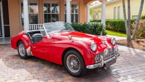 A red TR3A-spec 1960 Triumph TR3 by a peach-colored house