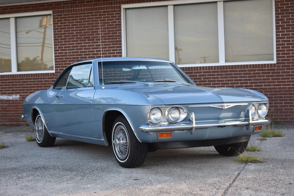 A light-blue 1965 Chevrolet Corvair Corsa by a red-brick building