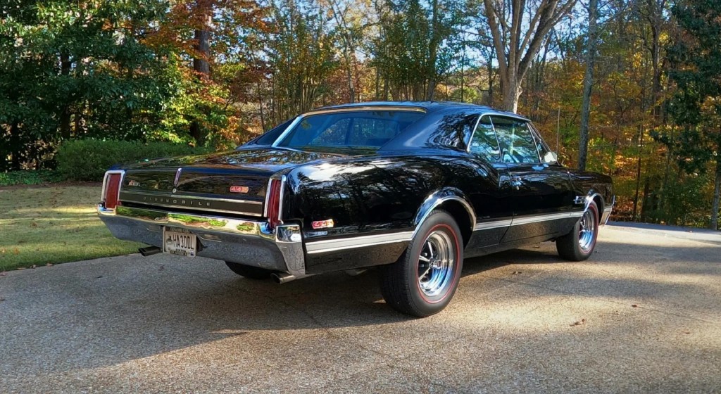 The rear 3/4 view of a black 1966 Oldsmobile 442 parked on a stone-lined driveway by some trees