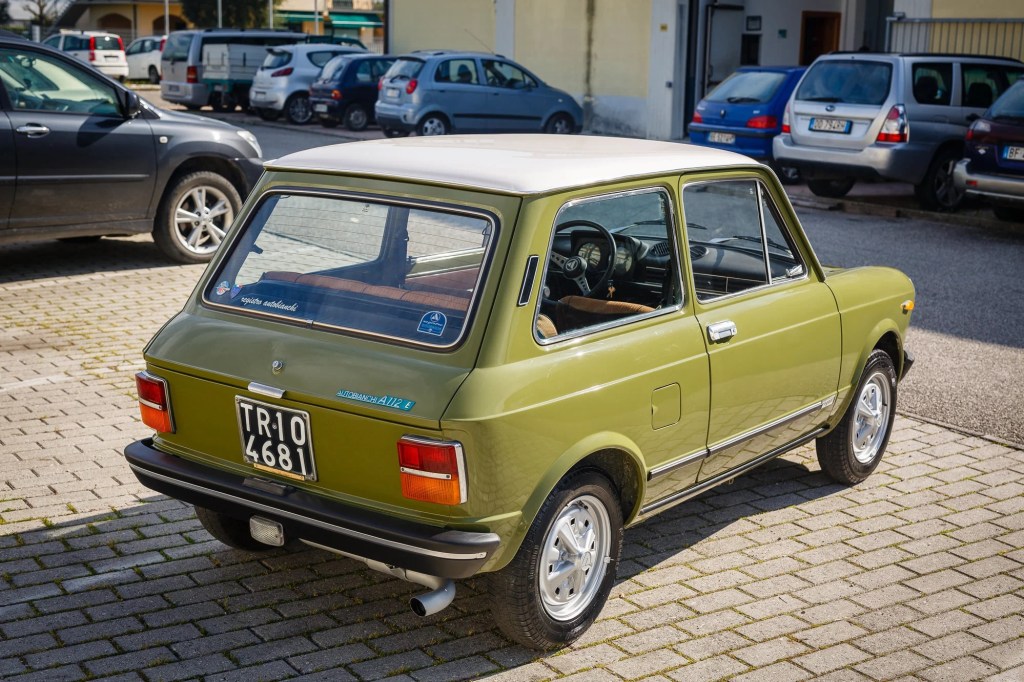 The rear 3/4 view of a green 1971 Autobianchi A112 Abarth in a crowded parking lot