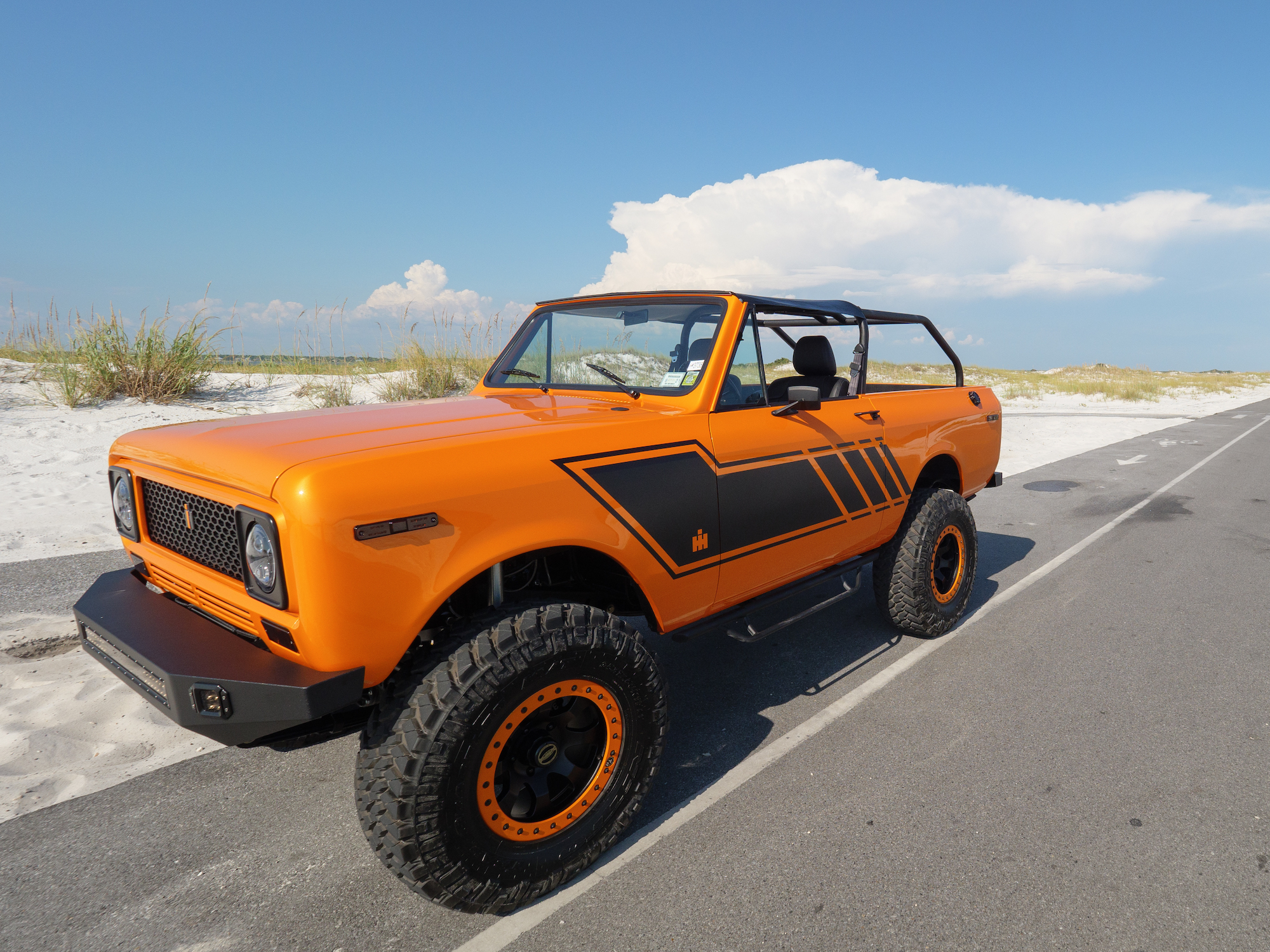 An orange 1979 International Harvester Scout SUV-like vehicle parked on the shoulder of a road next to a sand dune
