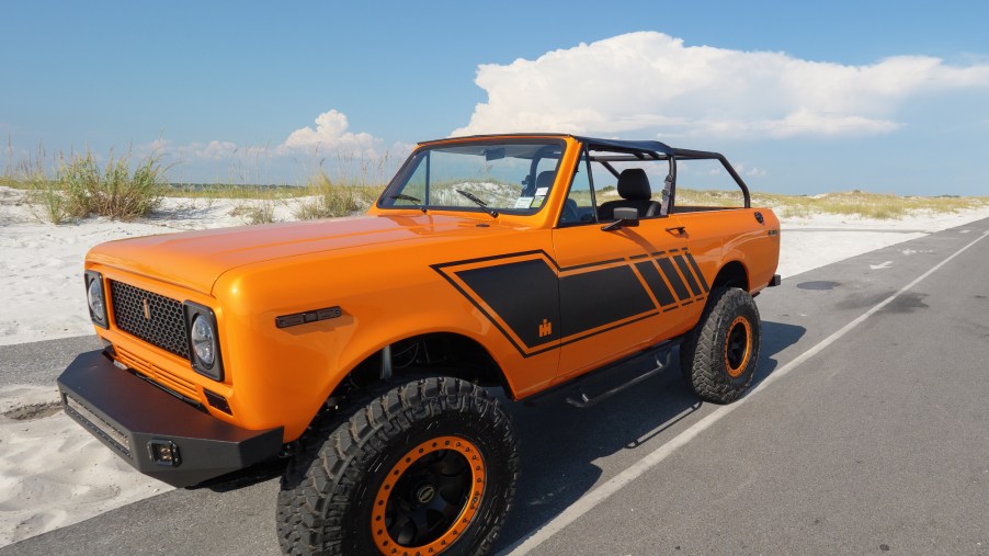 An orange 1979 International Harvester Scout SUV-like vehicle parked on the shoulder of a road next to a sand dune