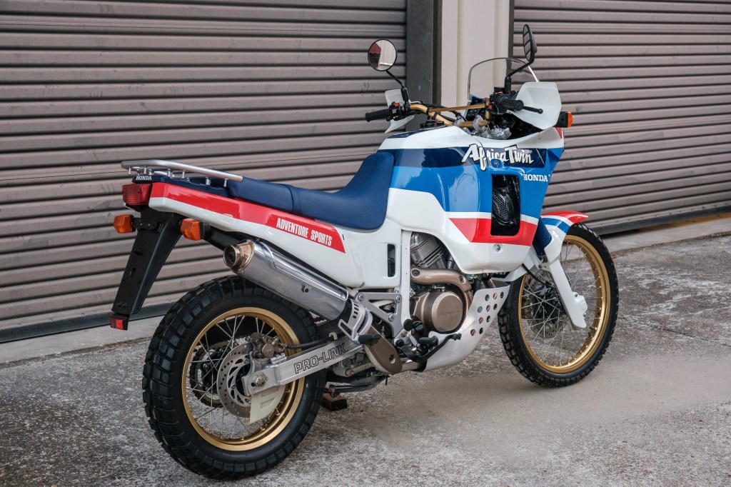 The rear 3/4 view of a white-blue-and-red 1989 Honda Africa Twin XRV650 by a garage