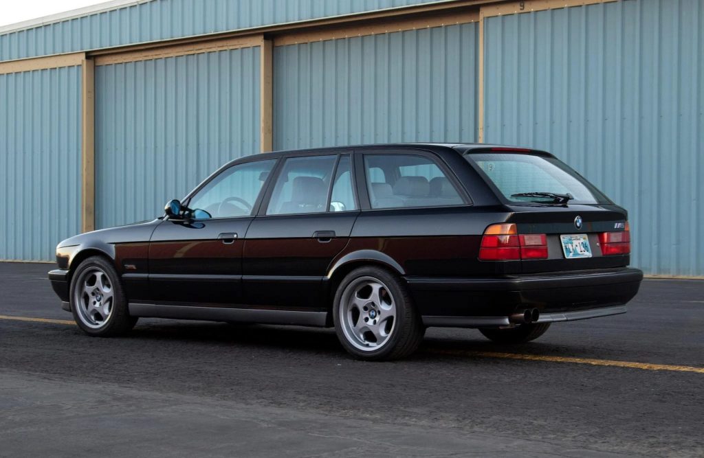 The rear 3/4 view of a black 1992 BMW M5 Touring by a blue warehouse