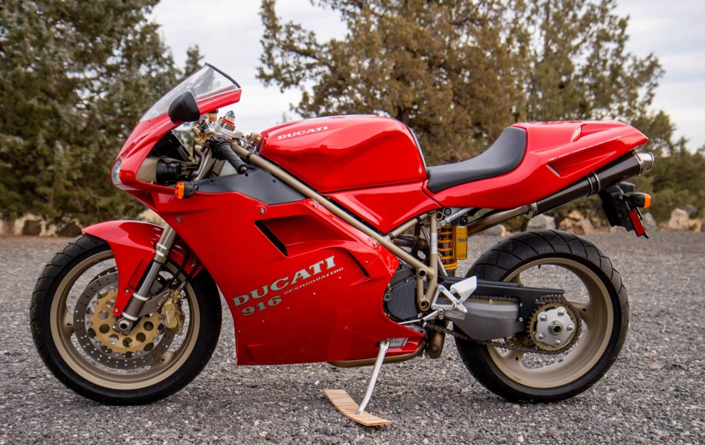 The side view of a red 1997 Ducati 916 parked on a gravel area by a forest