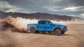 A blue 2019 Ford F-150 Raptor four-door pickup truck kicks up dust as it drives on dirt in front of a mountain on a cloudy day