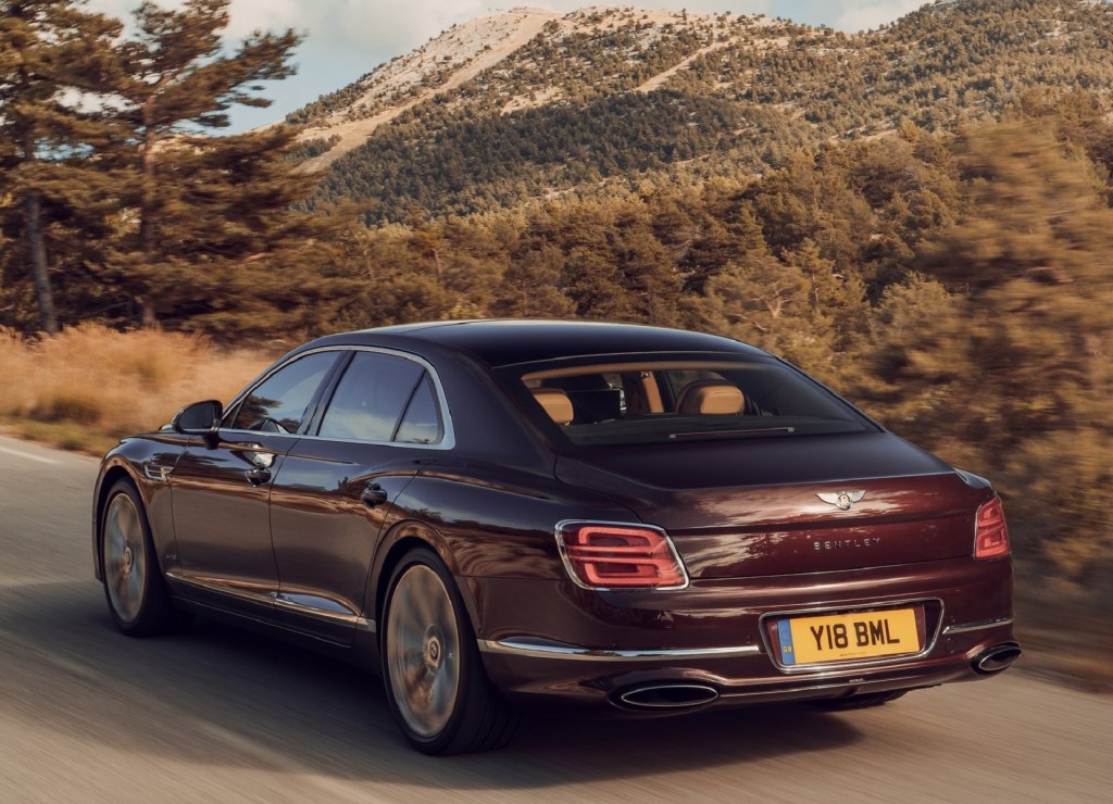 The rear 3/4 view of a maroon 2020 Bentley Flying Spur driving on a tree-lined mountain road at sunset