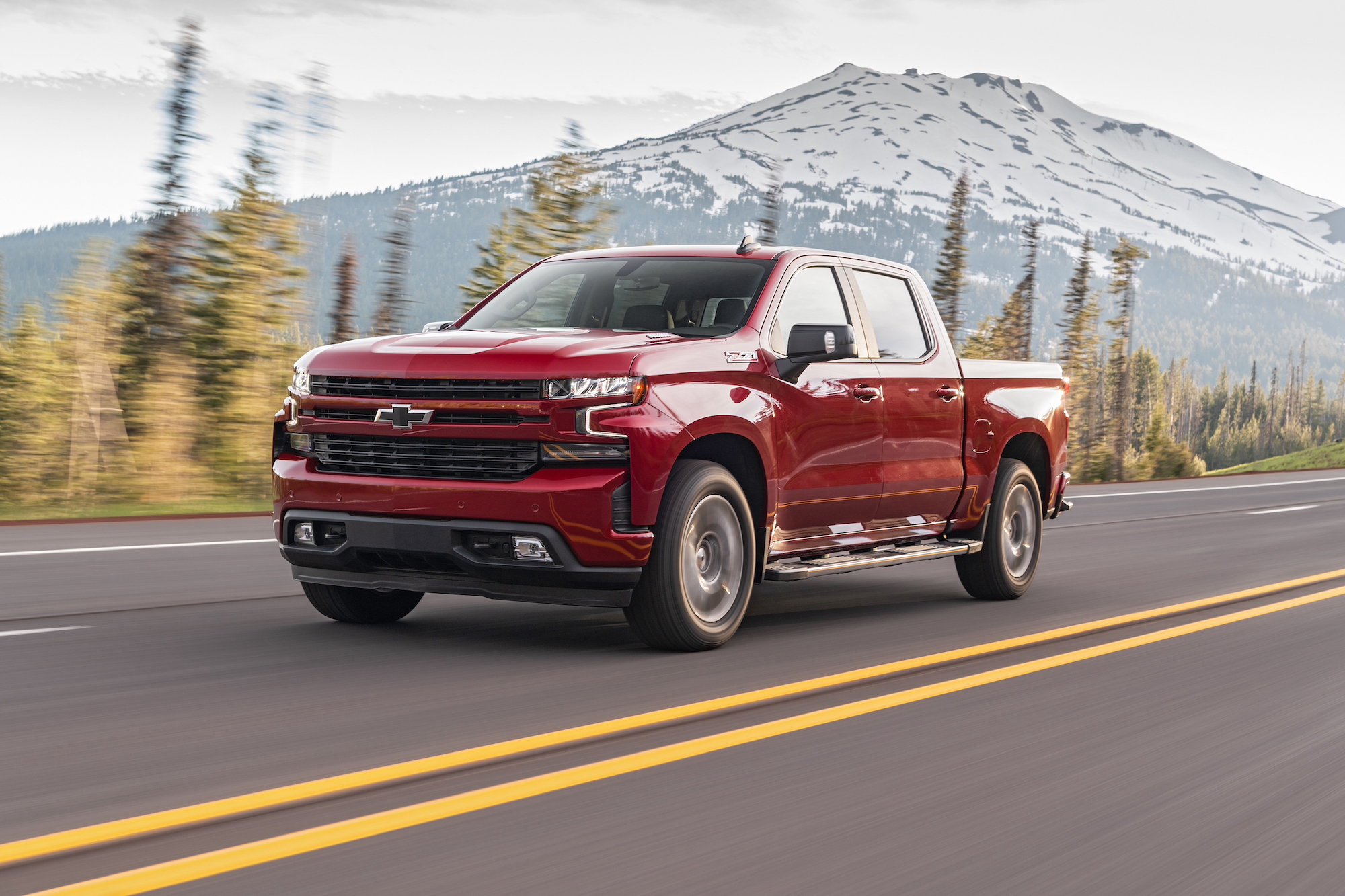 A red 2020 Chevrolet Silverado Diesel full-size pickup truck travels on a four-lane highway past pine trees and a snow-capped mountain