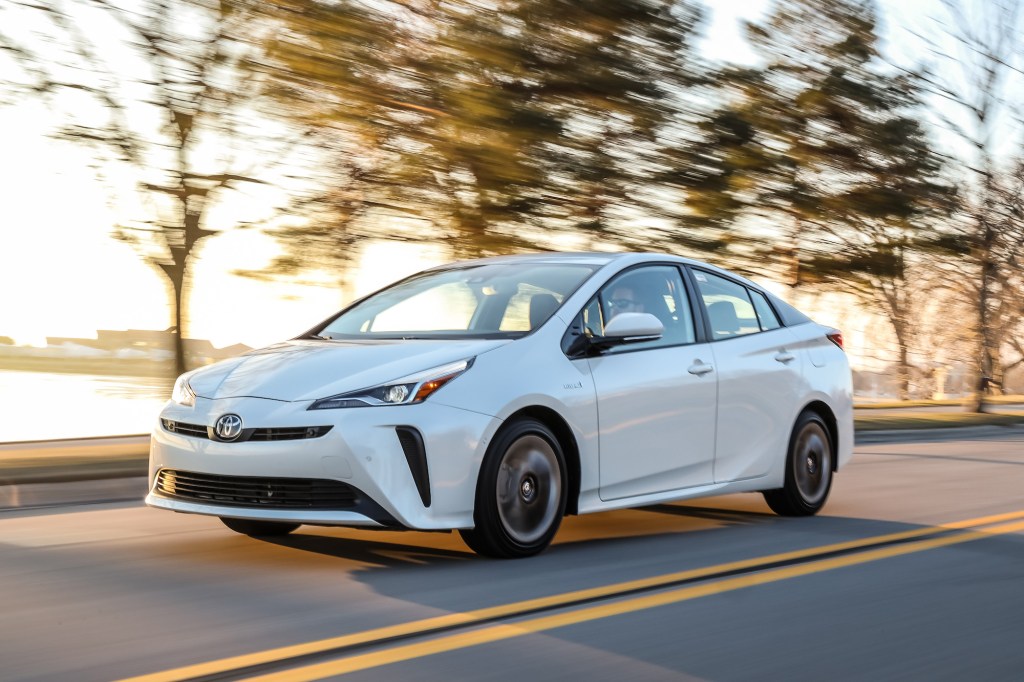 A white 2020 Toyota Prius, a Consumer Reports Green Choice, travels on a two-lane highway lined with trees and a body of water