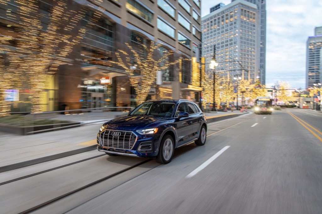 A dark-blue 2021 Audi Q5 compact luxury SUV travels on a city street lined with office buildings and leafless trees strung with glowing twinkle lights at dusk