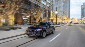 A dark-blue 2021 Audi Q5 compact luxury SUV travels on a city street lined with office buildings and leafless trees strung with glowing twinkle lights at dusk