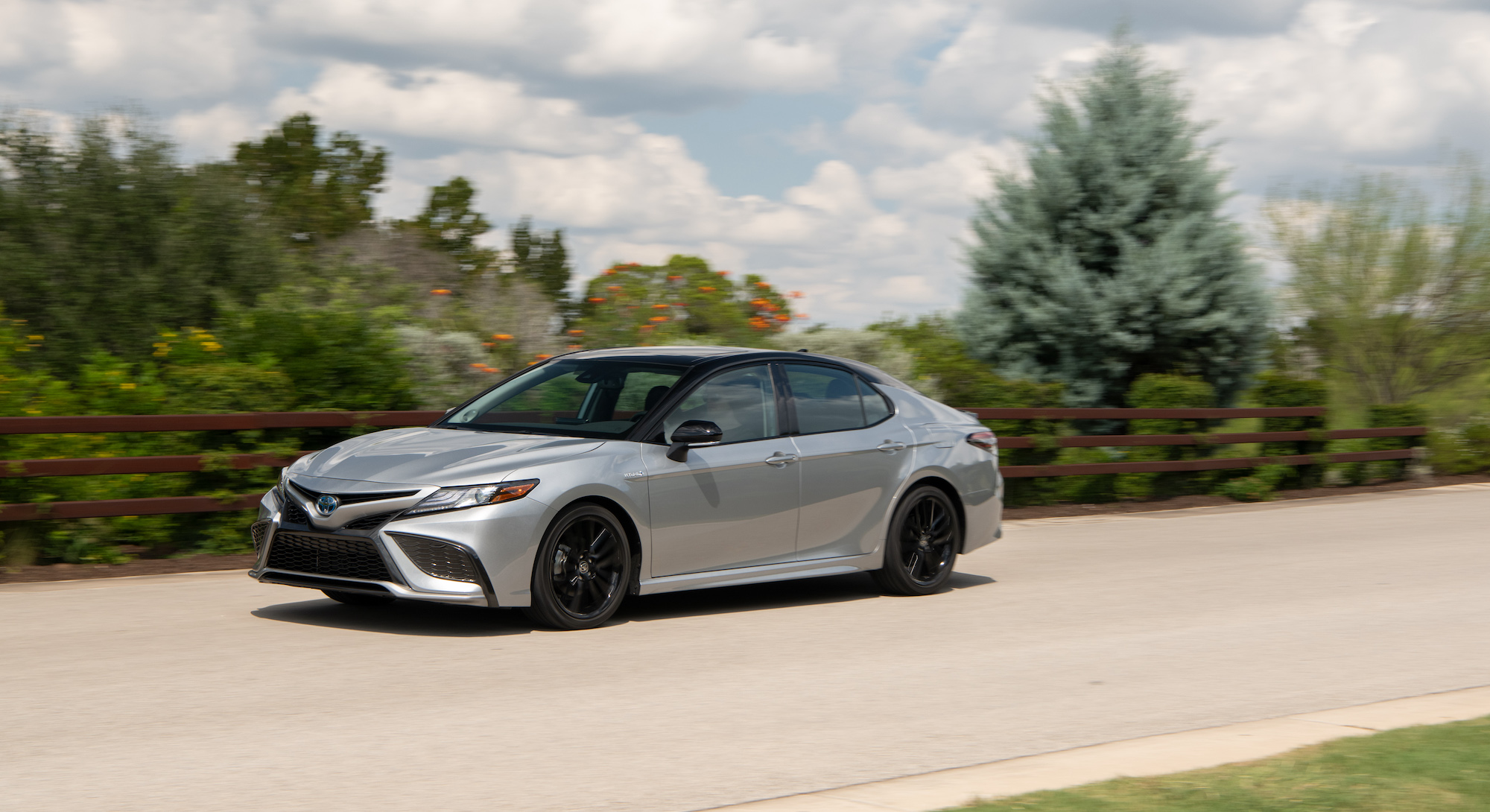 A silver 2021 Camry XSE Hybrid midsize sedan traveling on a country road along a brown wooden fence and foliage